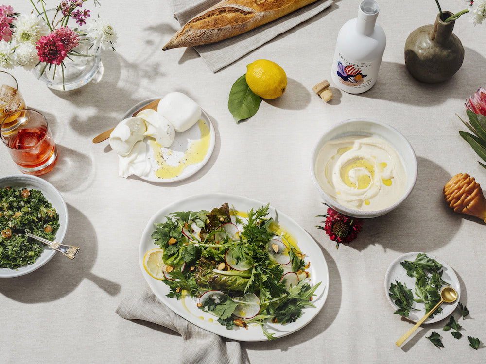 Image of tablescape with a dressed green salad, yogurt dip, burrata, floral arrangements, bread and cocktails.